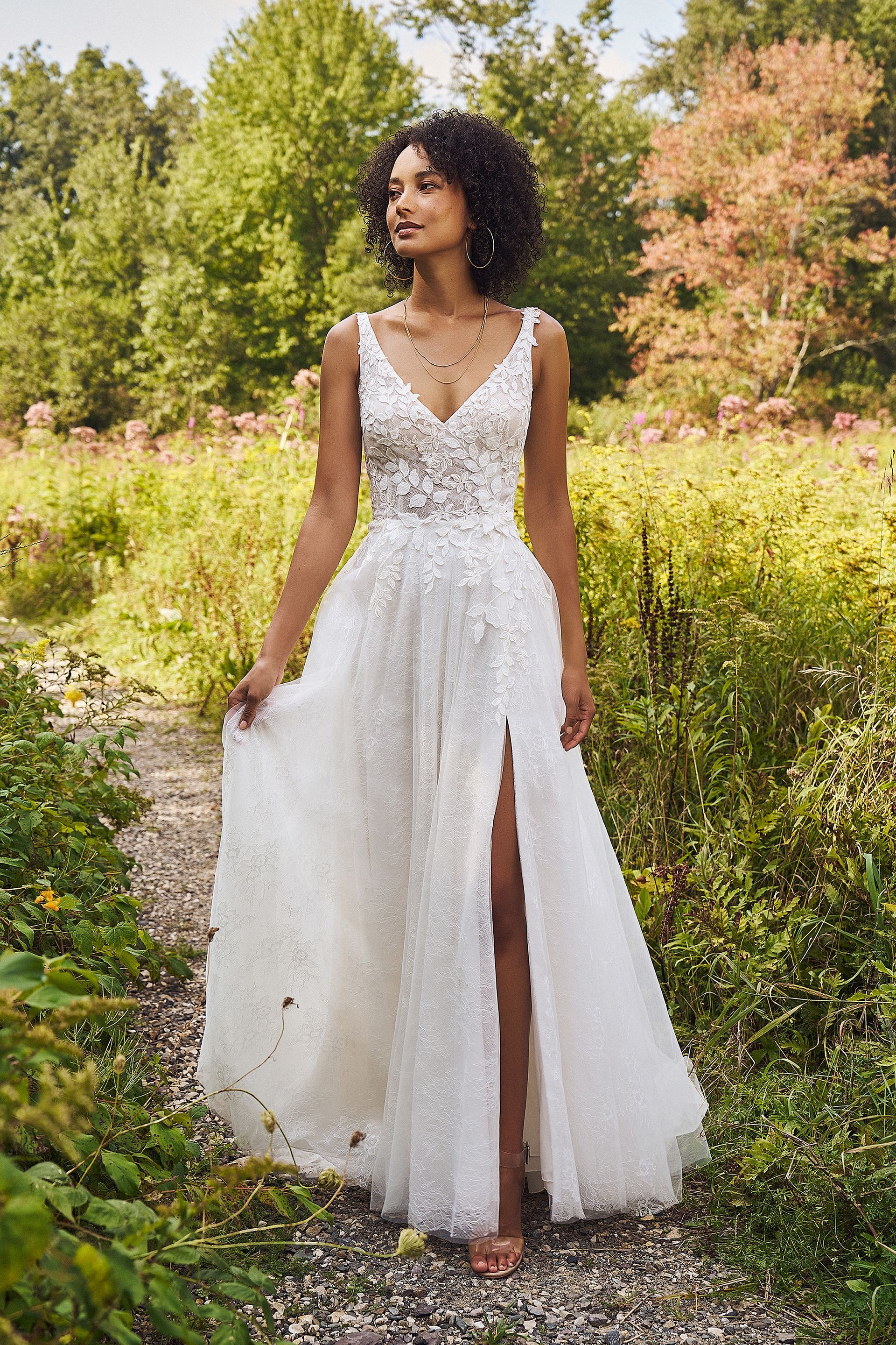 Woman in white wedding dress with a slit, standing in a sunny outdoor setting with trees.