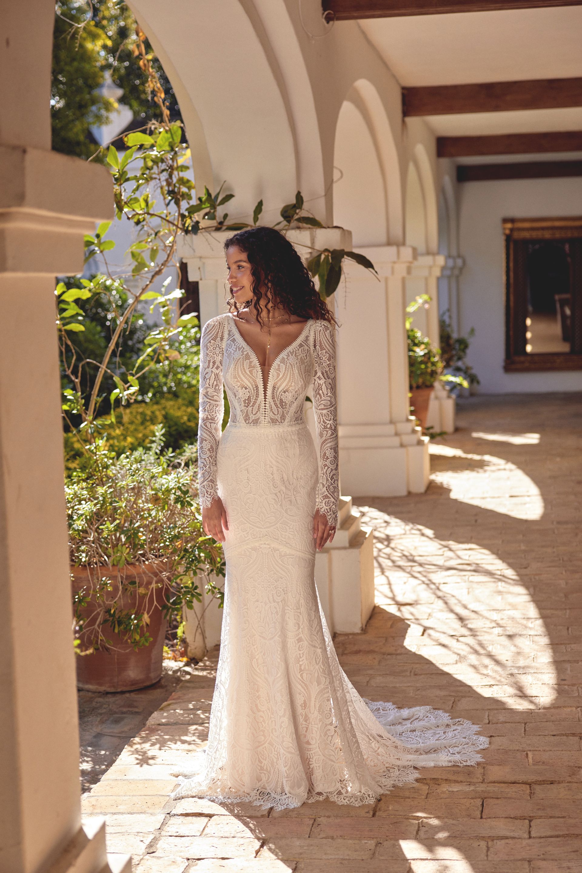 Woman in an off-the-shoulder, floral lace wedding gown, standing outdoors in a garden setting.