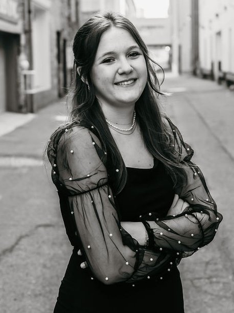 A woman in a black dress is standing in an alleyway with her arms crossed and smiling.