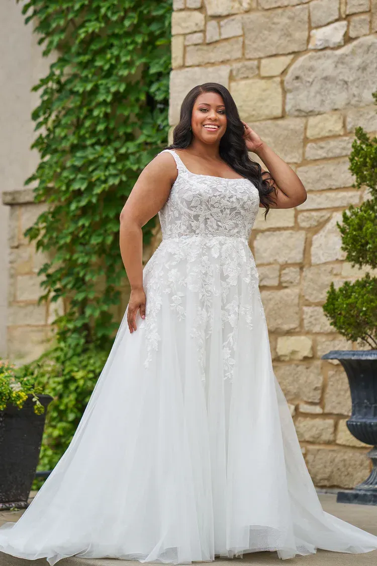 Woman in floral off-the-shoulder gown smiles, arms raised. Standing outside.