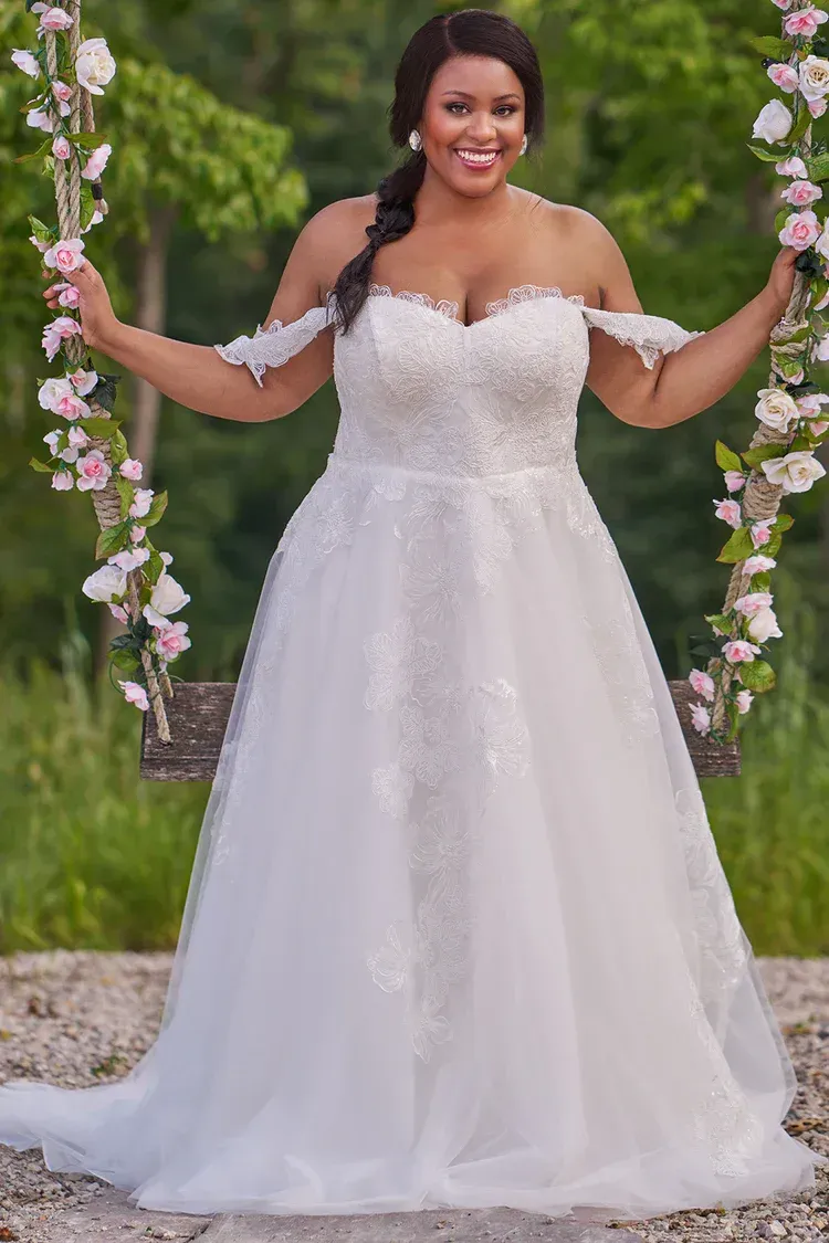 Woman in white off-the-shoulder wedding dress smiles, standing on a swing decorated with flowers.