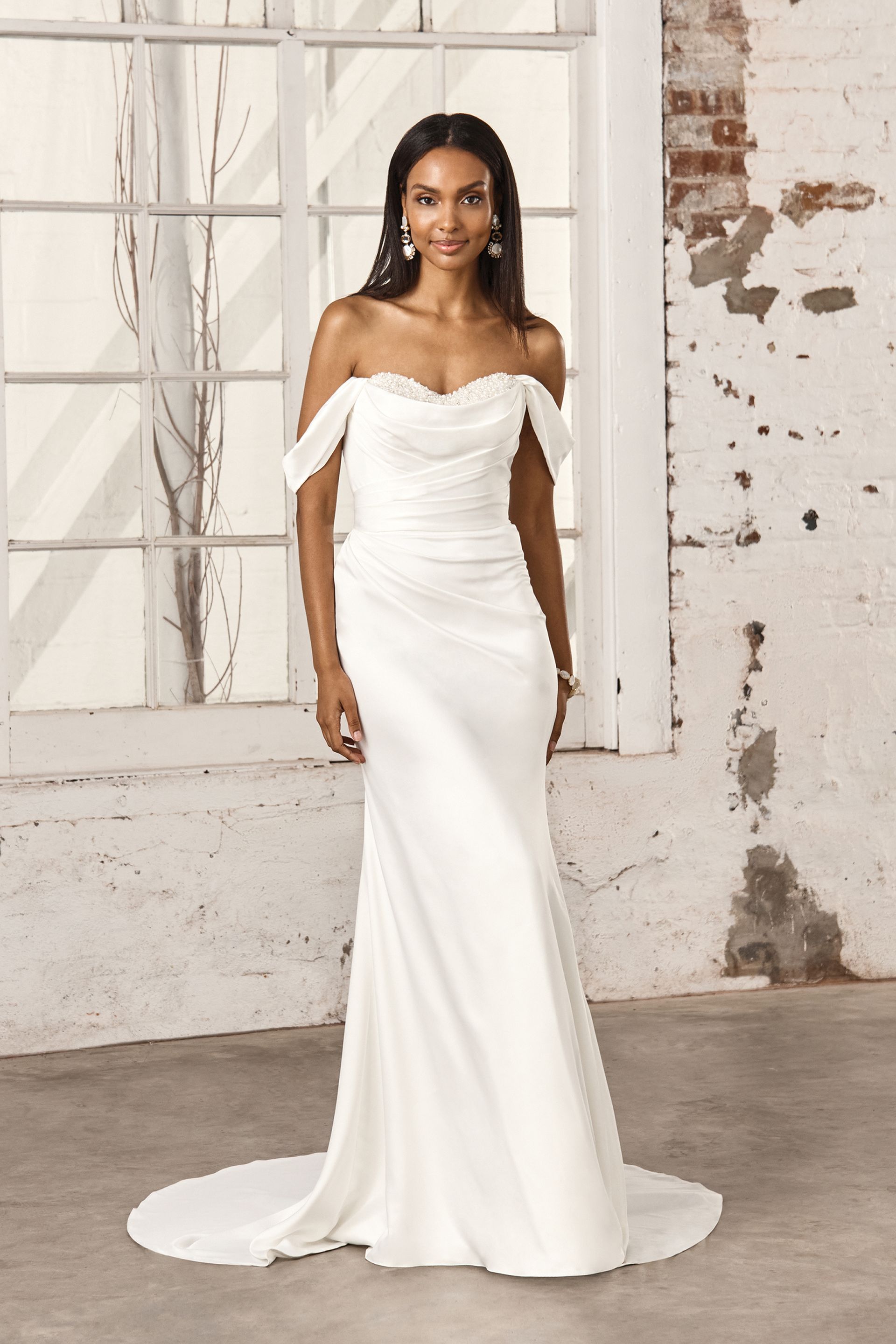 Woman in an off-white, off-the-shoulder wedding dress posing in front of a window and brick wall.