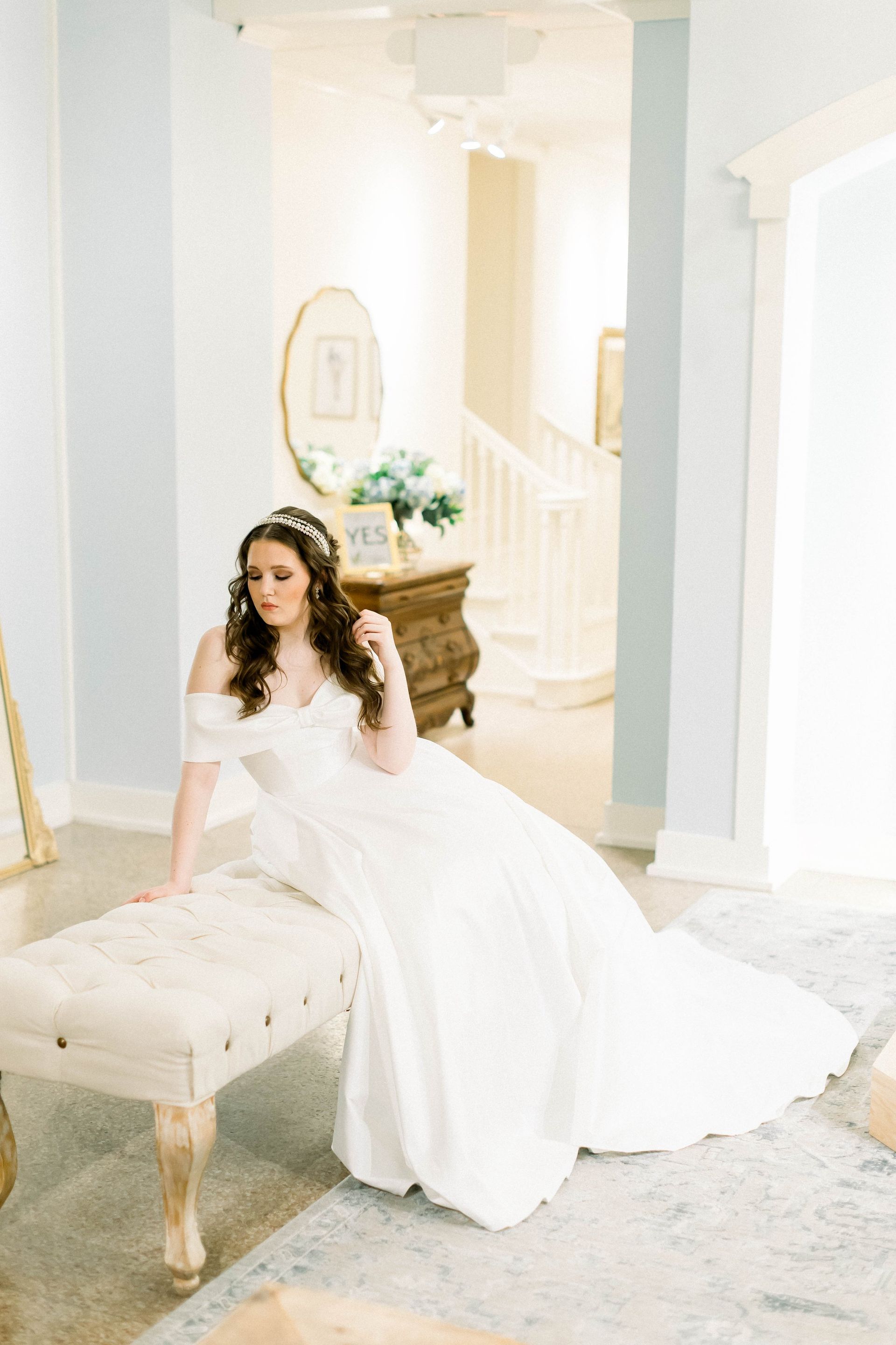Bride in a white off-the-shoulder gown sits on a cream bench in a light-filled room. She wears a tiara and has wavy brown hair.