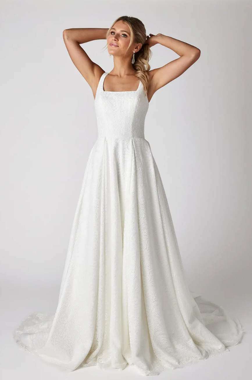 A woman in a white wedding dress is standing with her hands behind her head.