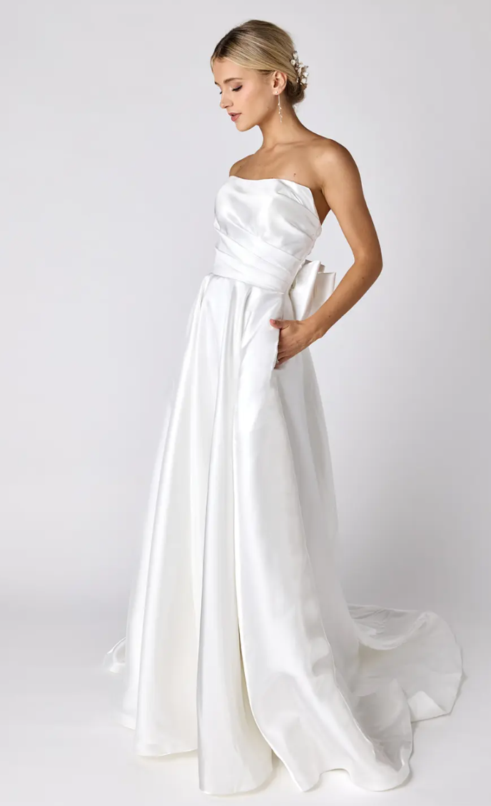 A woman in a white wedding dress is standing with her hands in her pockets.