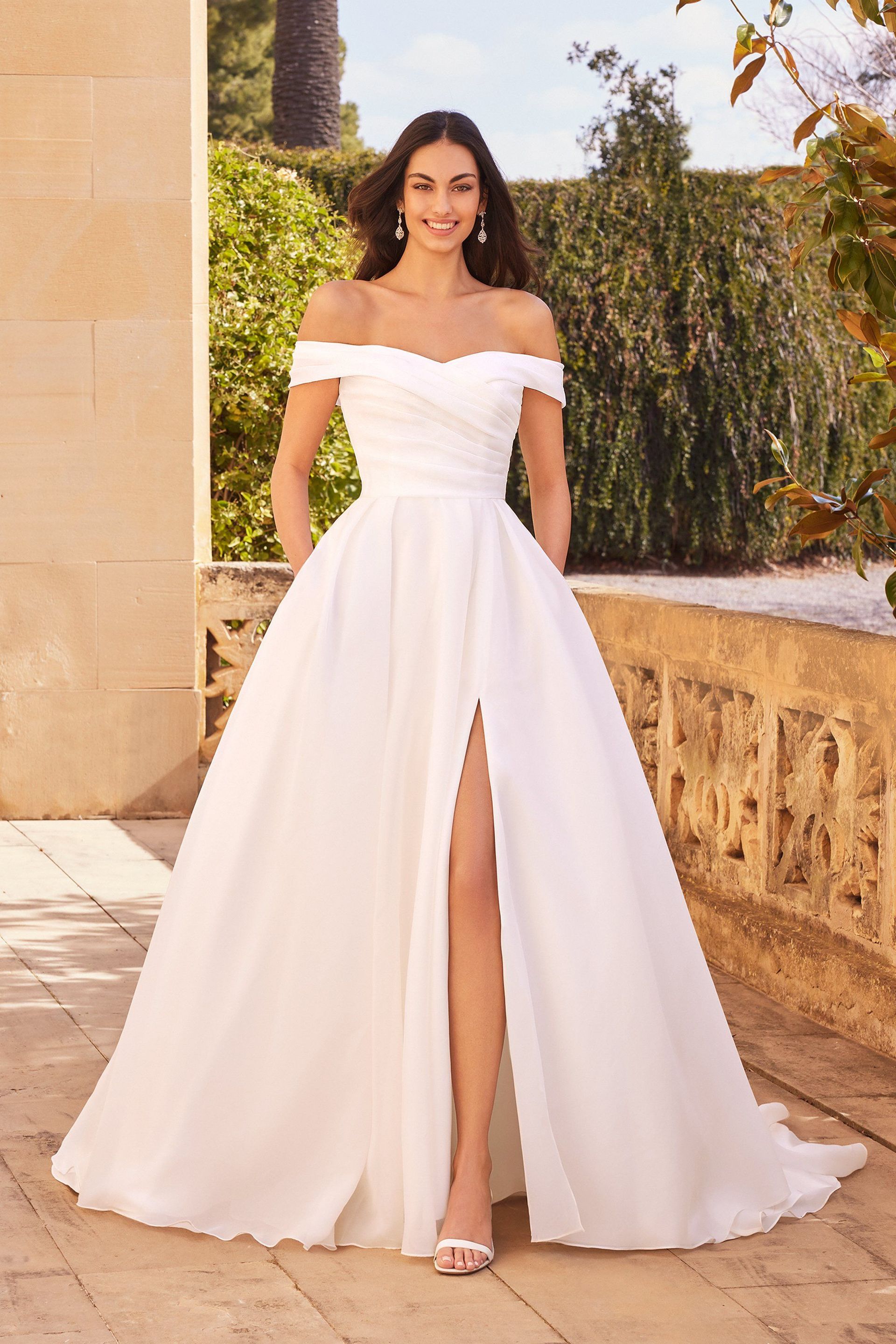 A woman is wearing a white off the shoulder wedding dress with a high slit.