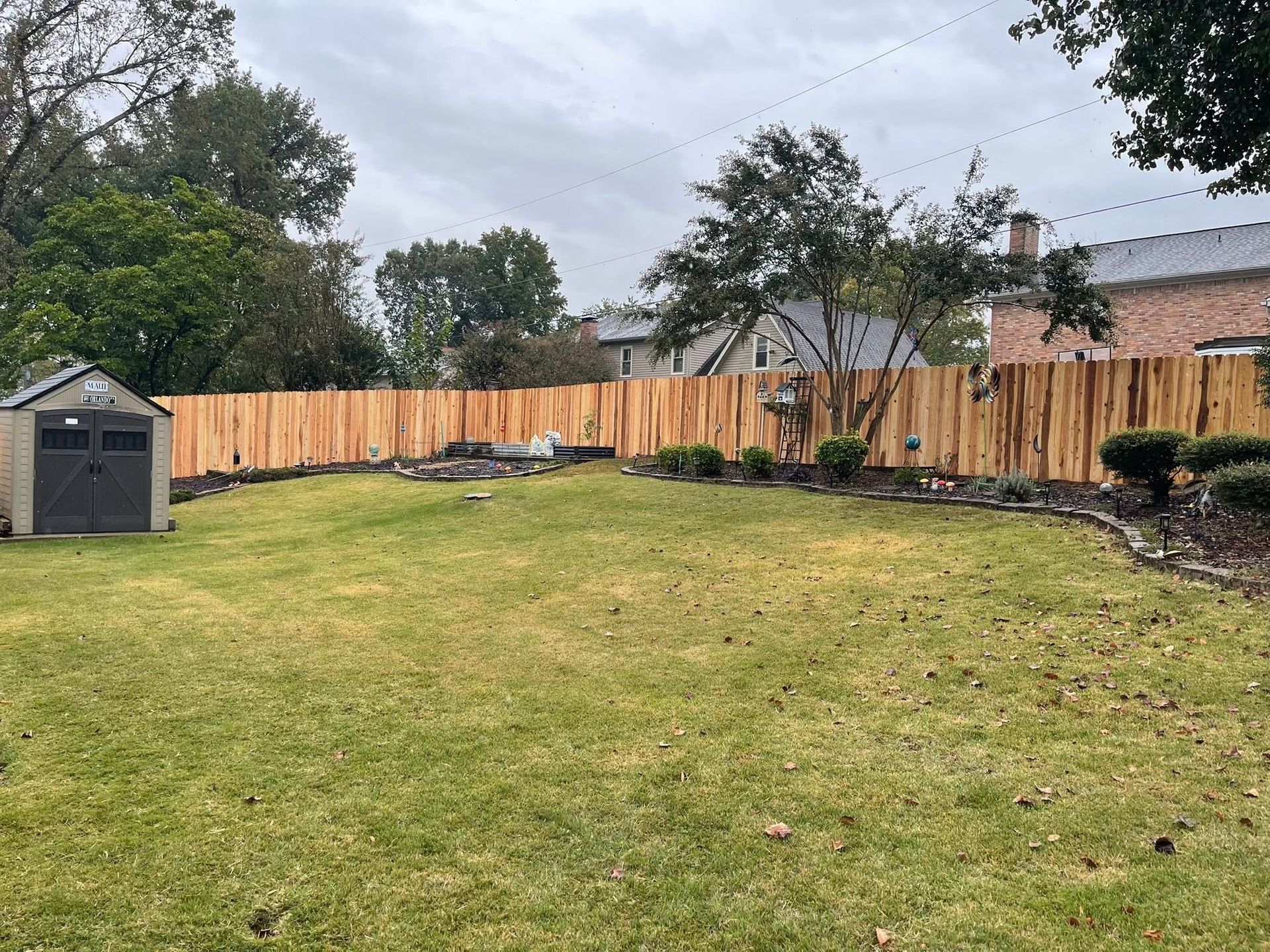 Backyard with a green lawn, wooden fence, shed, and small garden beds under an overcast sky