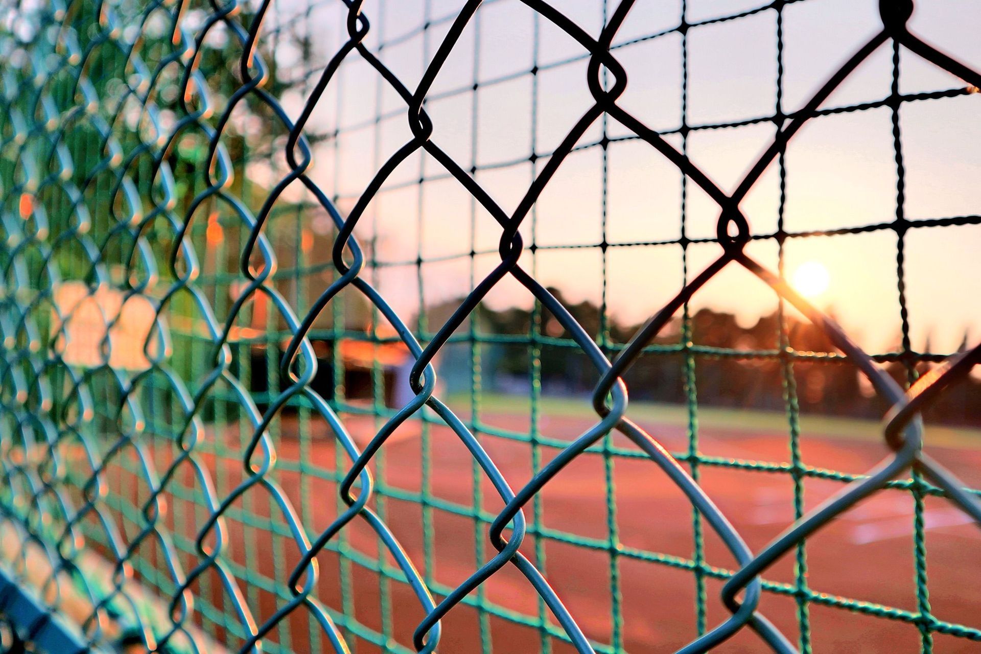 Chain-link fence in front of a tennis court at sunset, with orange and green playing surface.