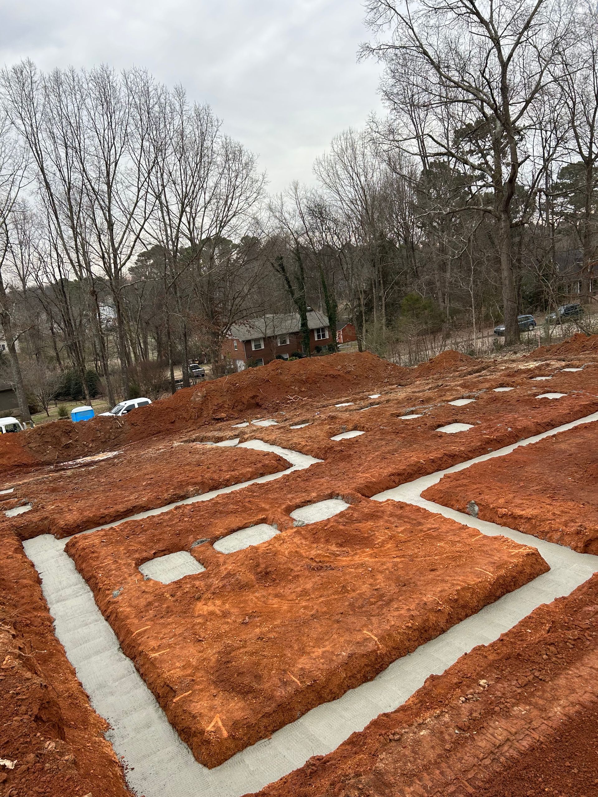 Foundation trenches dug in red soil, ready for concrete, with trees and a building in the background.