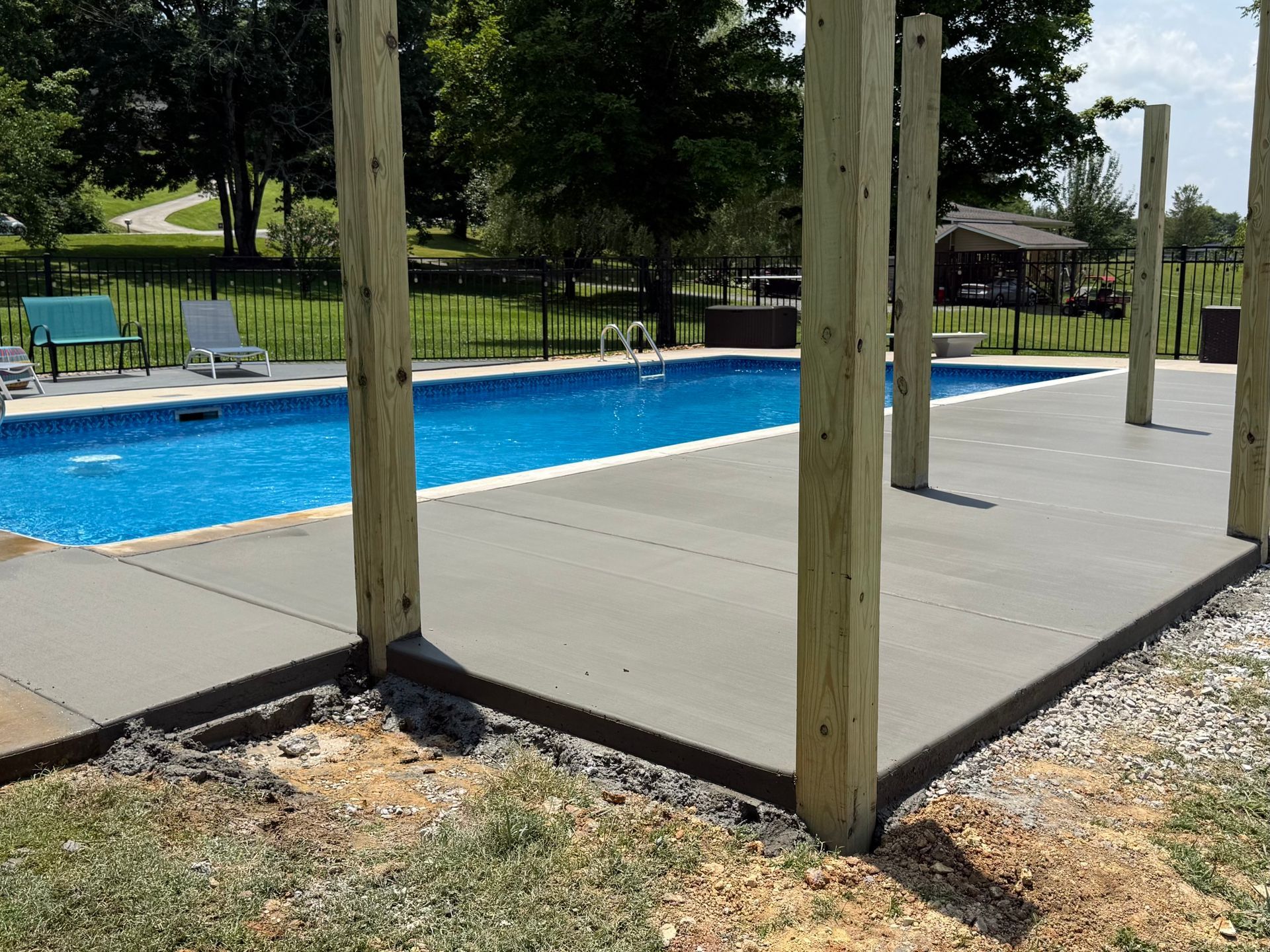 Newly poured concrete patio next to a swimming pool, with wooden support beams.