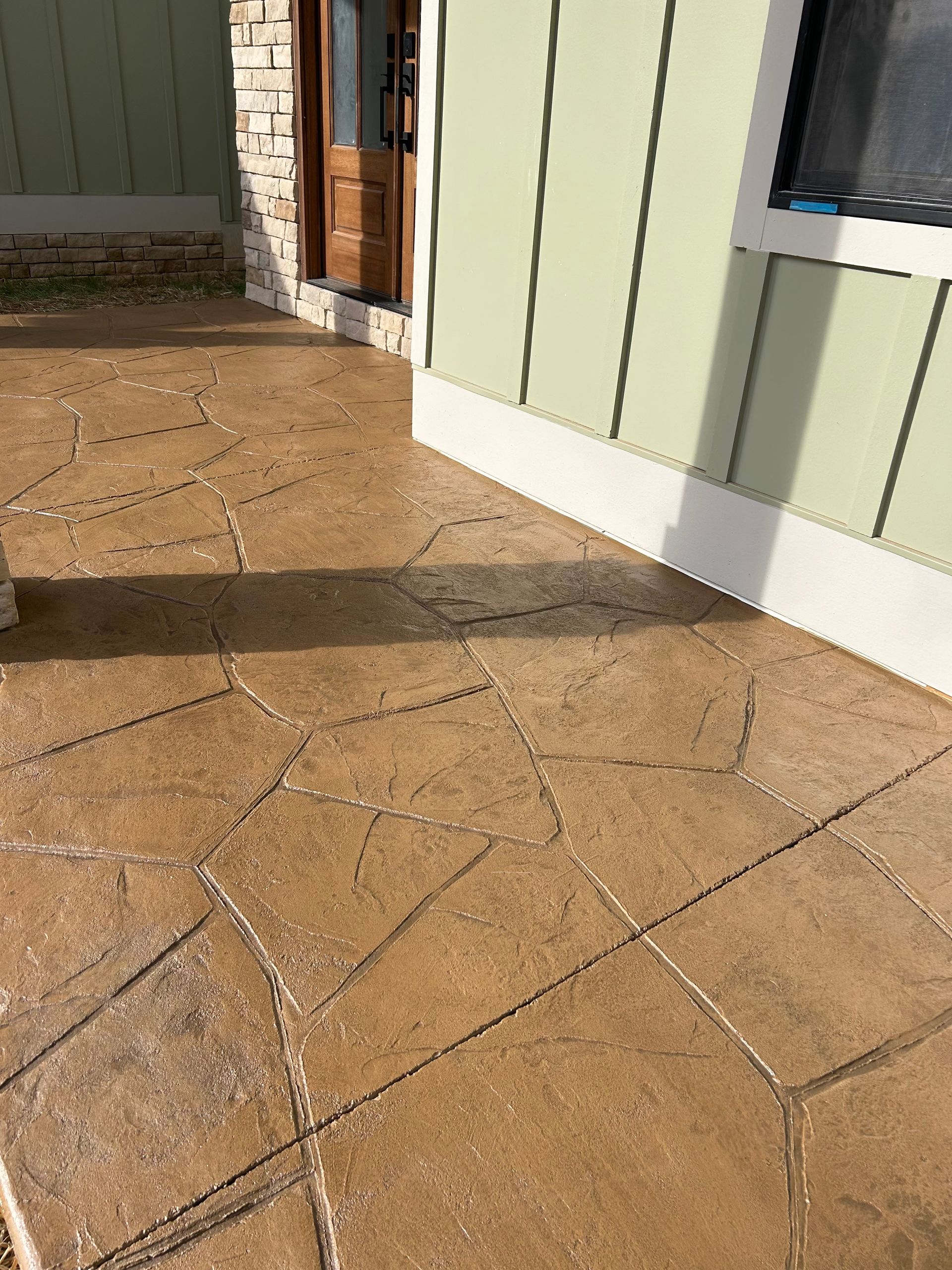 Tan stamped concrete patio next to a light green house with a brown door.