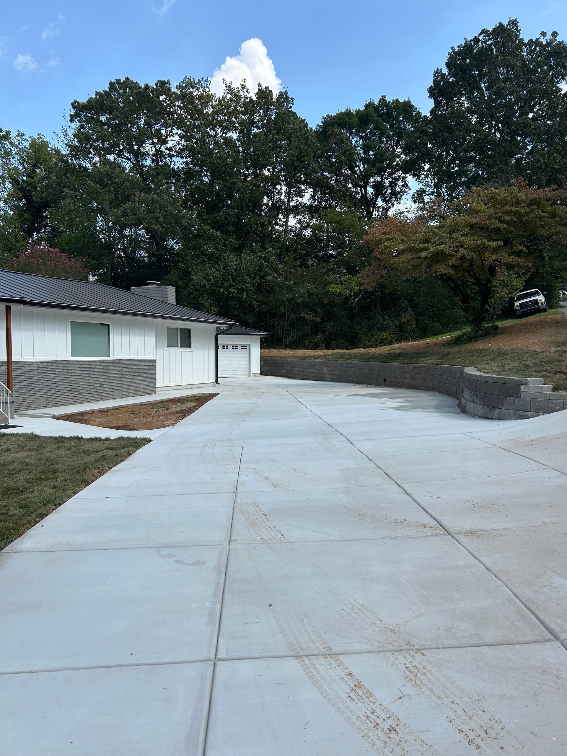 Concrete driveway leading to a white house with a garage, backed by trees under a partly cloudy sky.