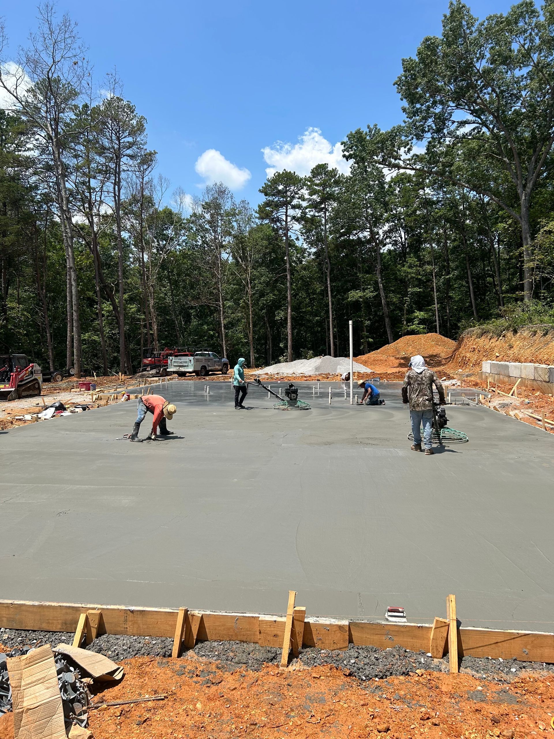Construction workers smoothing fresh concrete on a foundation, surrounded by trees under a blue sky.