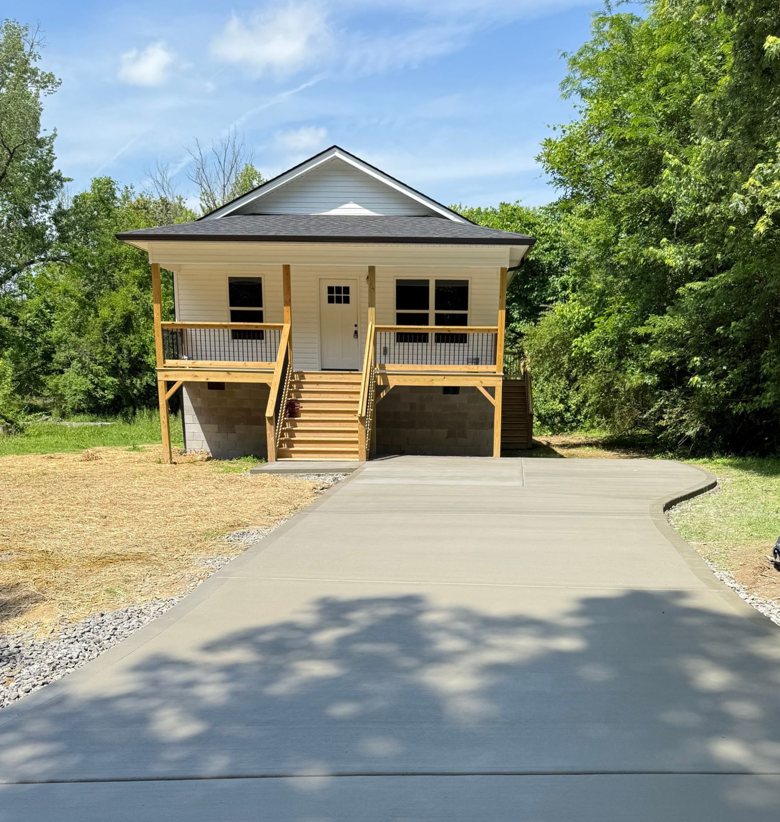 Small white house with a porch and wooden stairs. Concrete driveway leads to the house, surrounded by trees.