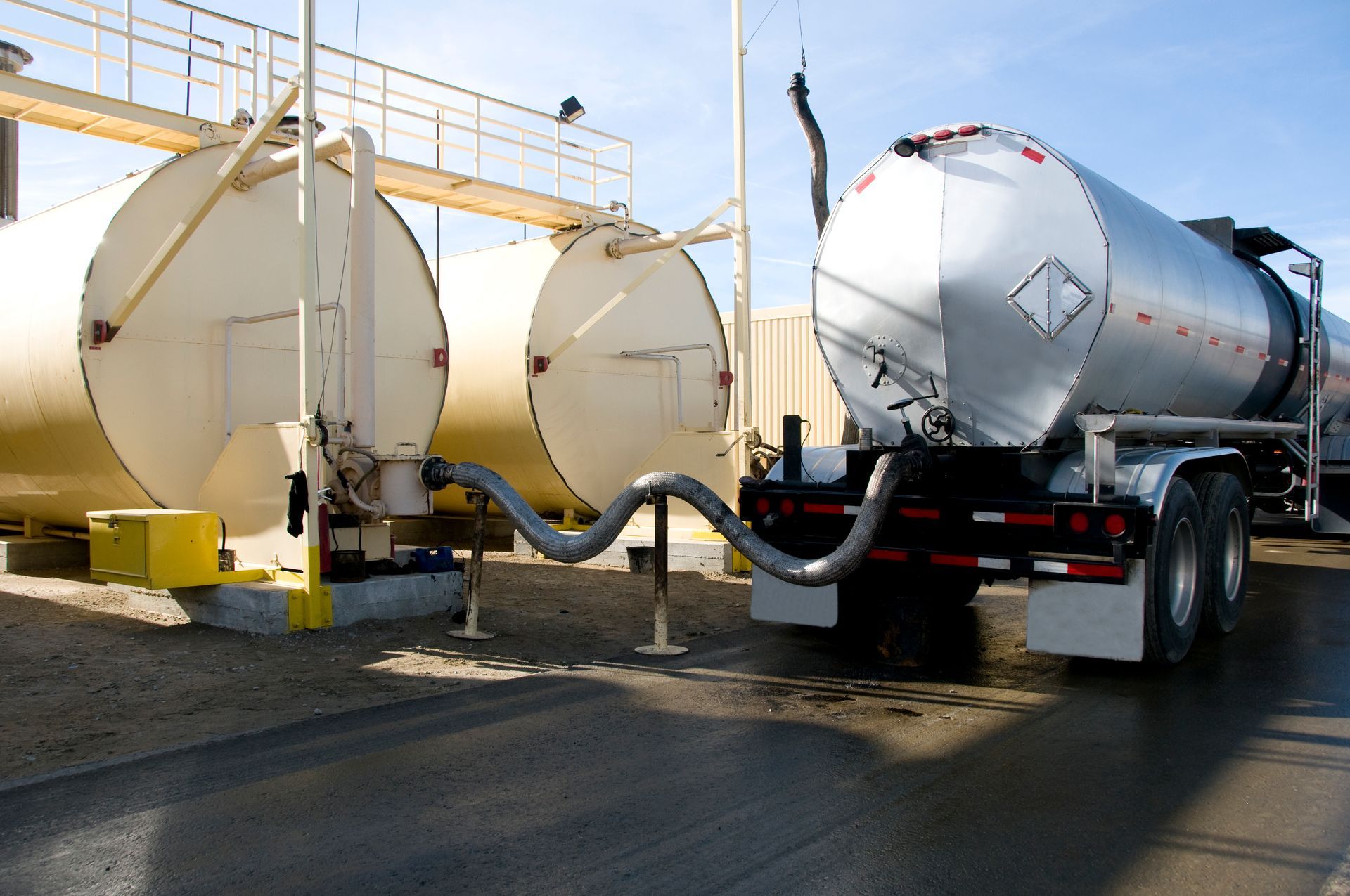 An Oil tanker truck is parked next to two large storage tanks, connected by a hose.