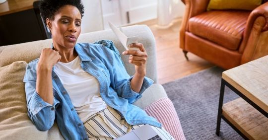 Woman sitting on couch, looking at a card, wearing blue shirt and striped pants, interior living room.