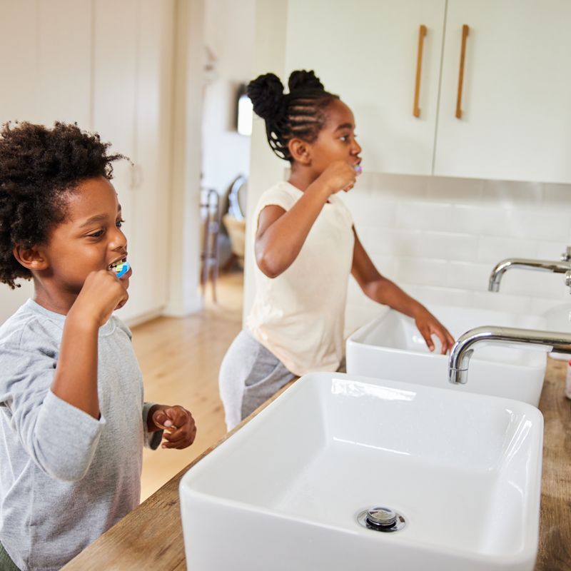 Two Black children brushing their teeth in a bathroom with white sinks and cabinets.
