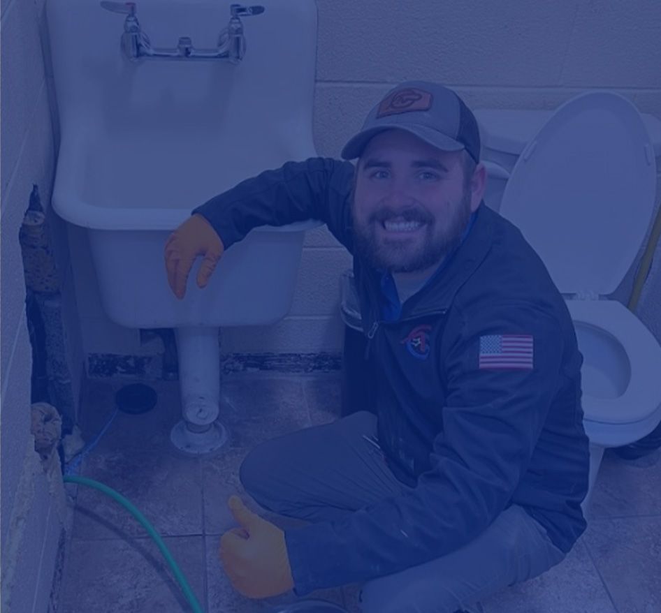 Plumber kneels near sink and toilet, giving a thumbs-up. Wearing cap and work clothes, smiling.
