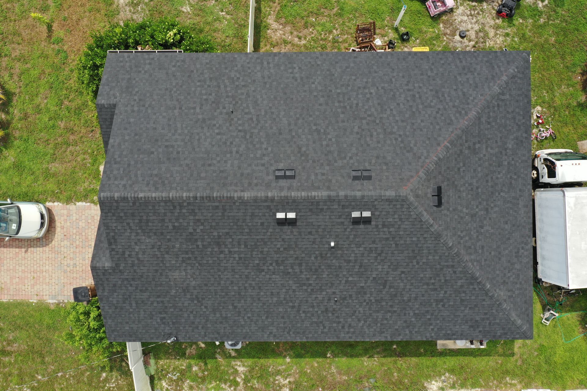 Aerial view of a dark gray shingle roof with three vents, surrounded by grass and a driveway.