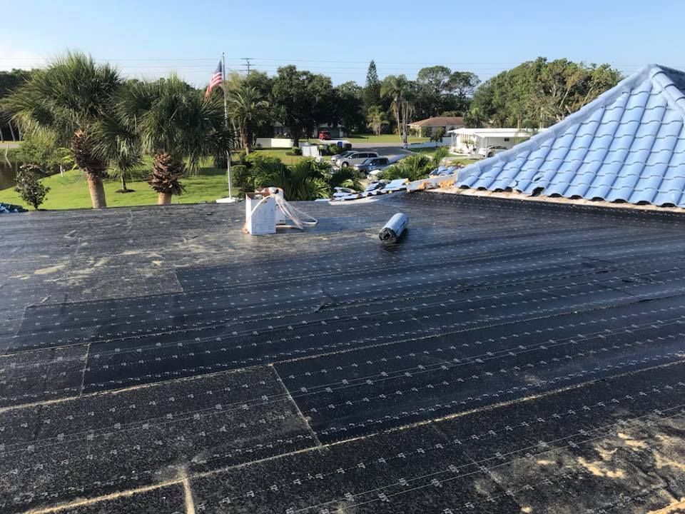 Rooftop with black roofing material partially installed; blue tarp on top, residential setting.