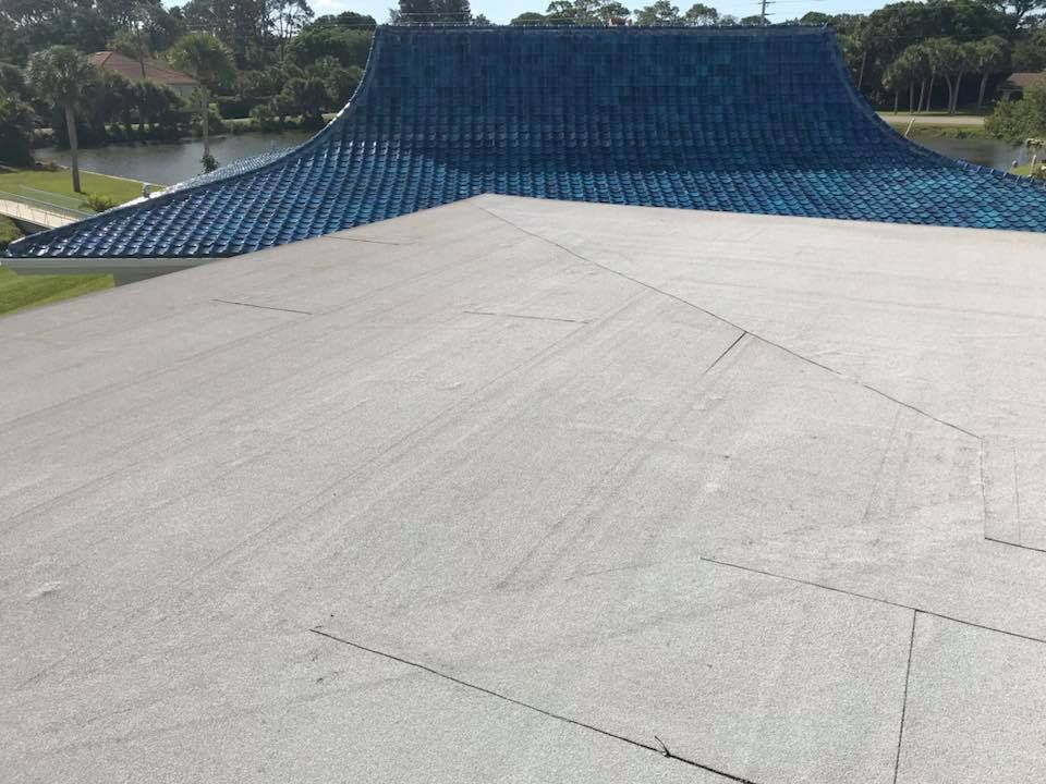 Flat, gray roof in foreground, with a blue tile roof in the background near a body of water.