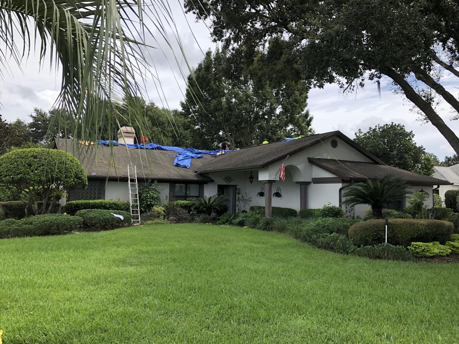 House with a roof partially covered in blue tarp; ladder leaning against side; lush green lawn and landscaping in front.