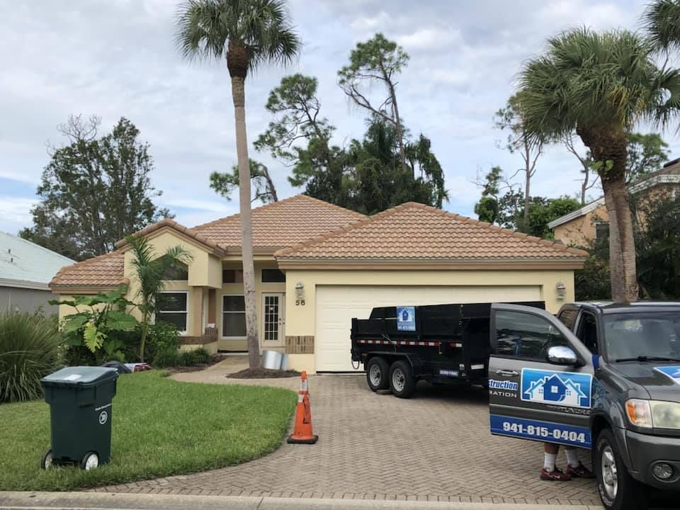 House with a brown tile roof and a dumpster trailer in the driveway, possibly for roofing work.