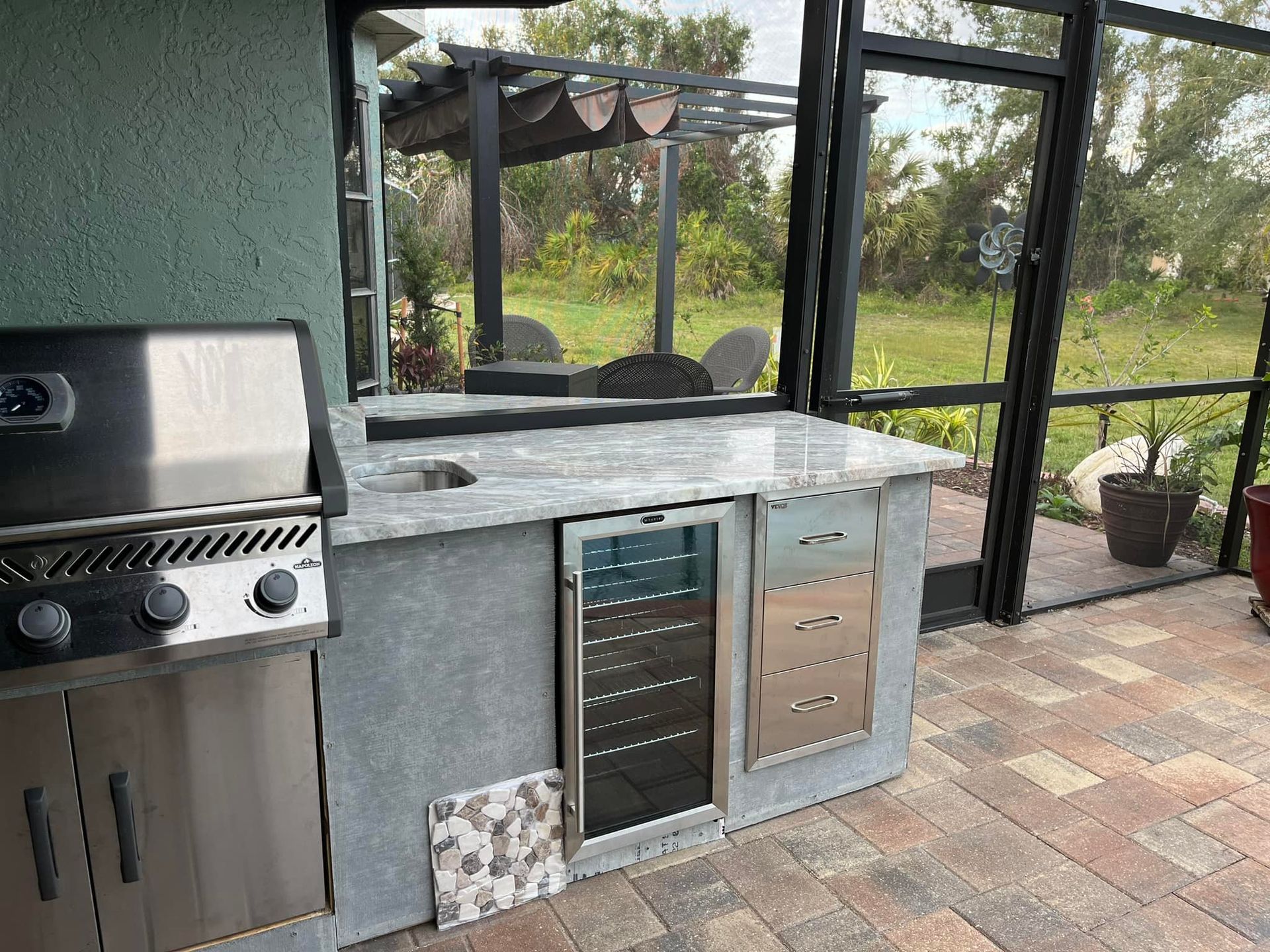 Outdoor kitchen with grill, counter, and appliances, adjacent to screened-in patio and backyard.