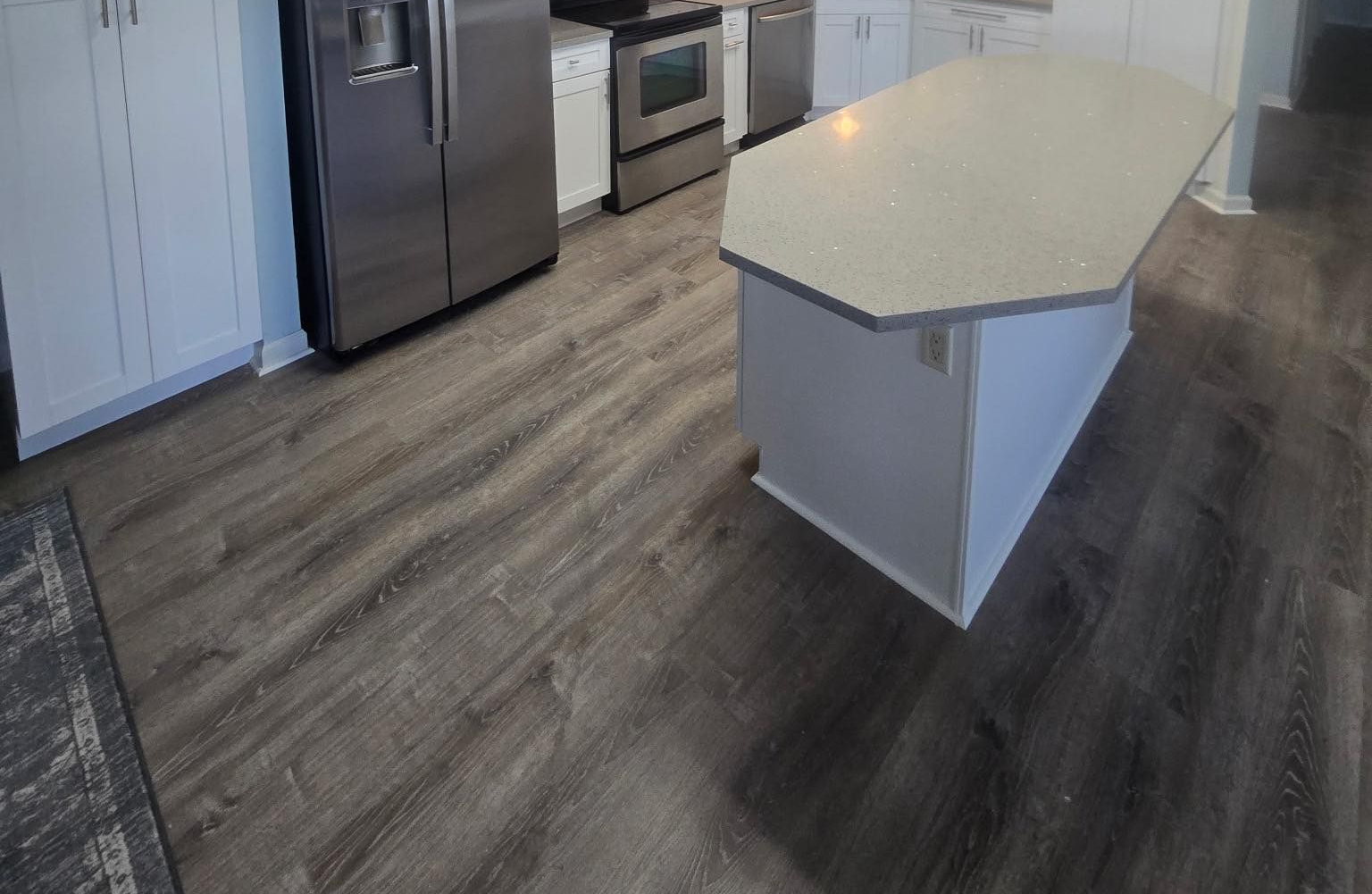 Kitchen with grey wood-look flooring, stainless steel appliances, white cabinets, and a white countertop island.