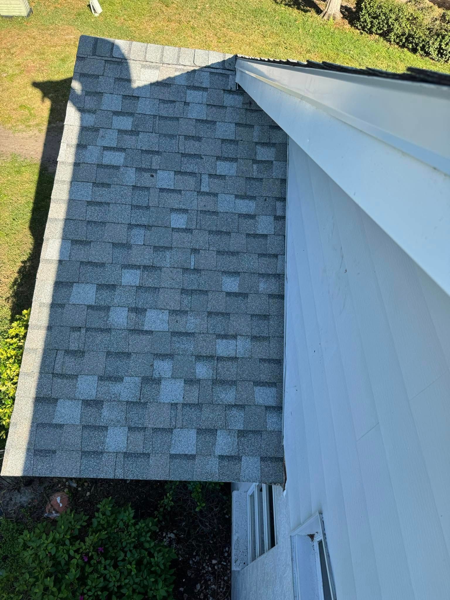 Gray shingle roof adjacent to a white wall. Green grass and foliage are visible in the background.