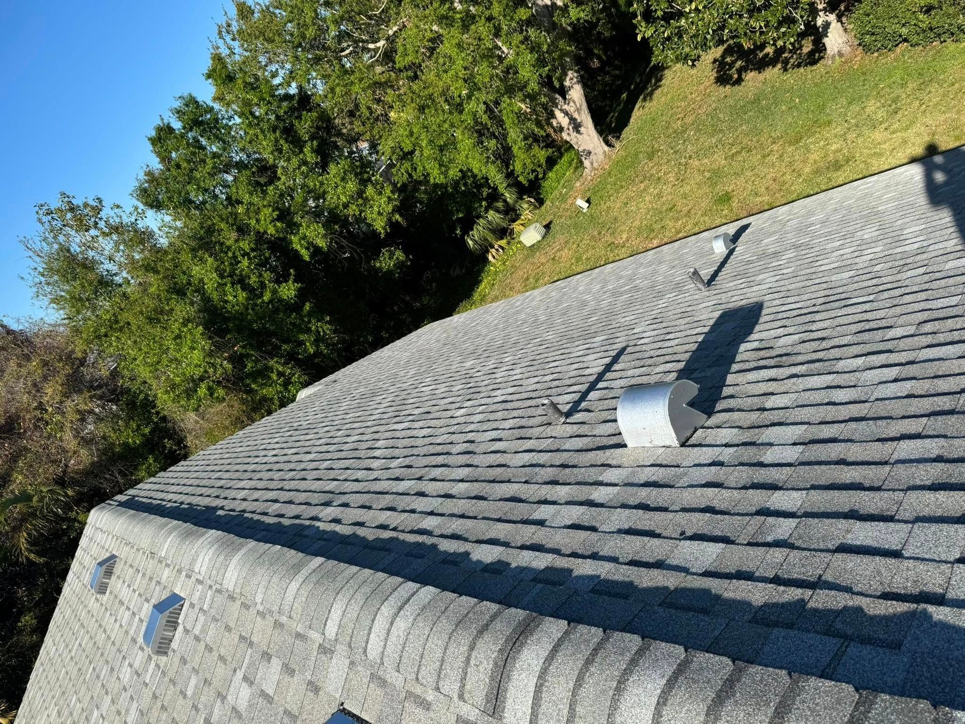 Gray asphalt shingle roof with several silver vents, set against a backdrop of green trees and a blue sky.