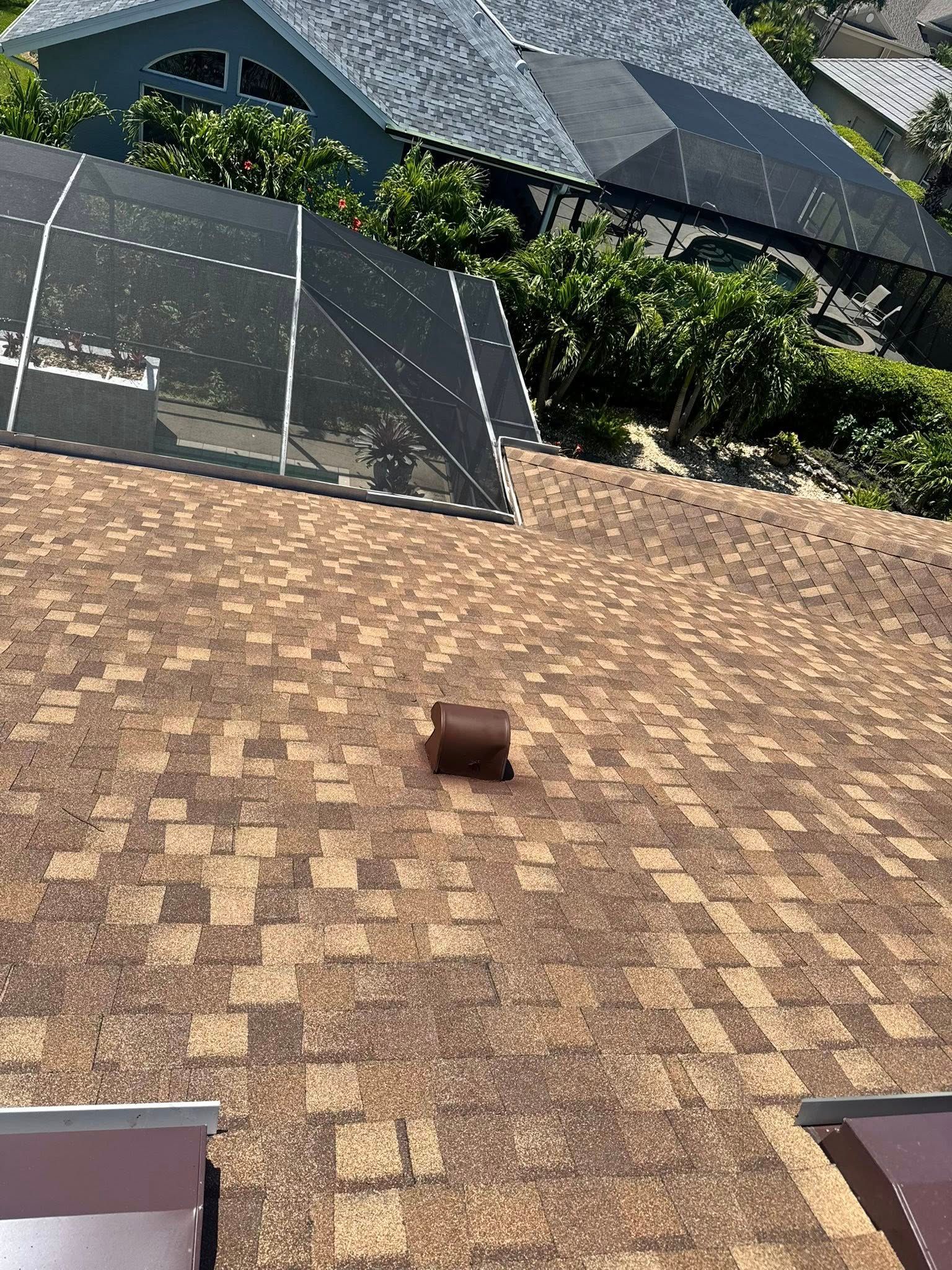 A brown shingled roof with a vent in the center, a screened-in pool enclosure, and another roof in the background.