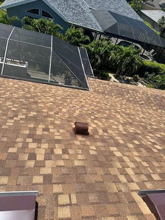 Brown shingled roof with a vent, surrounded by other roofs and lush greenery.