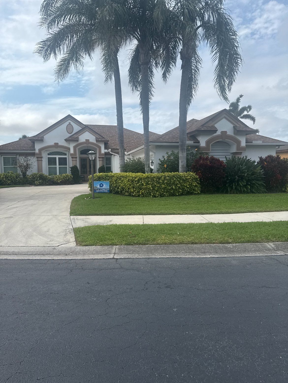A white stucco house with a curved driveway, palm trees, and green bushes.