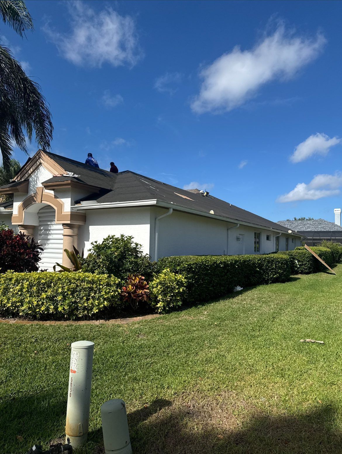 Roofers working on a black shingled roof of a white house, sunny day.