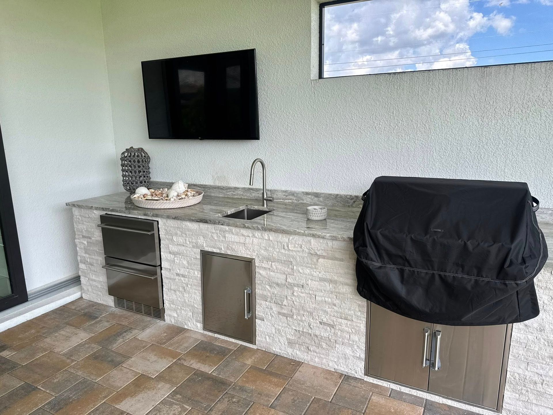 Outdoor kitchen with a grill, sink, refrigerator, and a TV mounted on a textured wall.