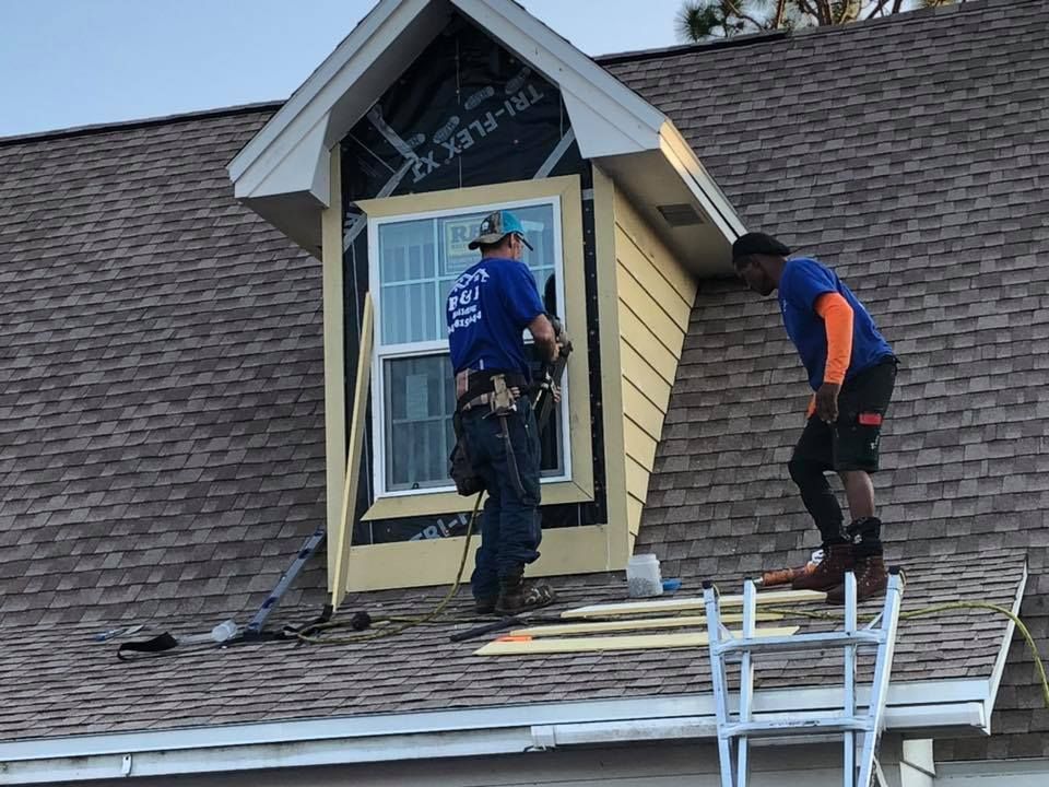 Two workers on a roof installing siding on a small dormer, sunny day.