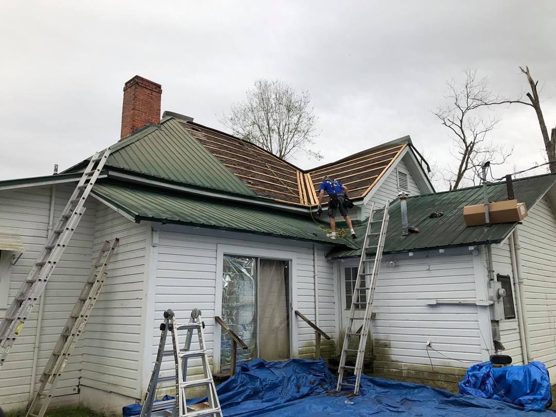 Person on roof, working on shingles and gutters. Ladders, white house, cloudy sky.