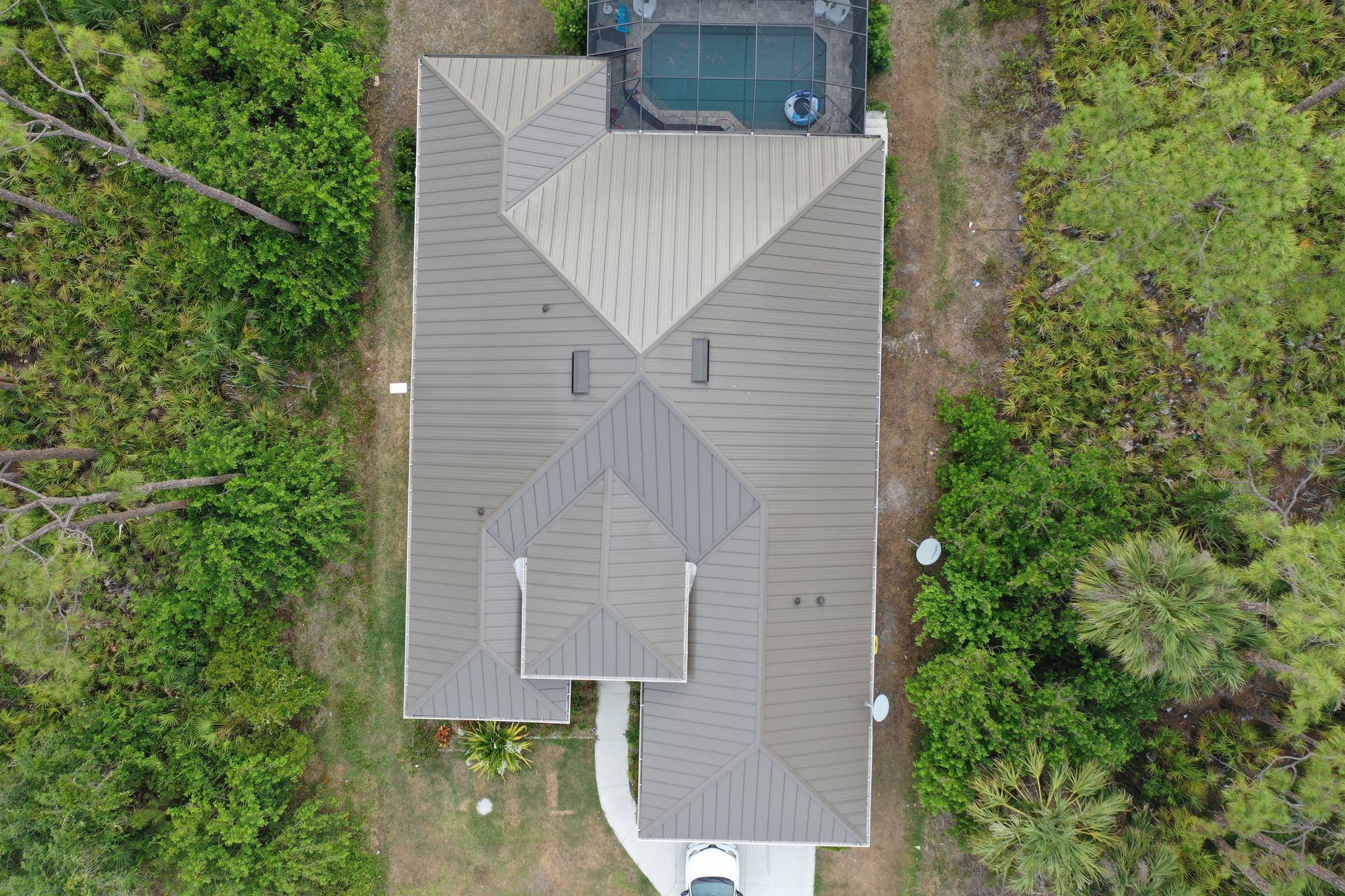 Overhead view of house with a pool, surrounded by green trees. Brown roof, white driveway.