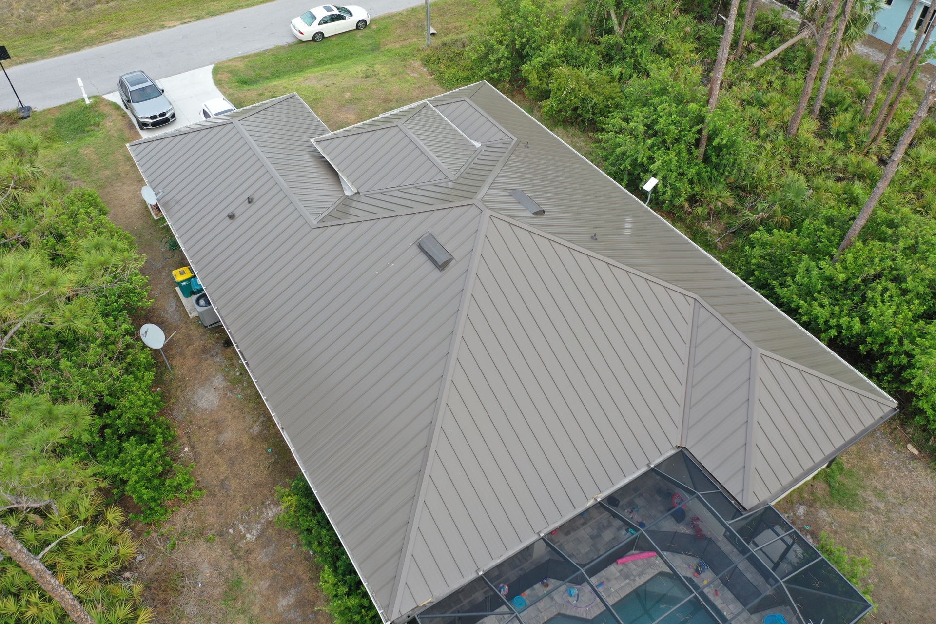 Aerial view of a house with a gray metal roof, surrounded by trees, a pool, and a driveway with cars.