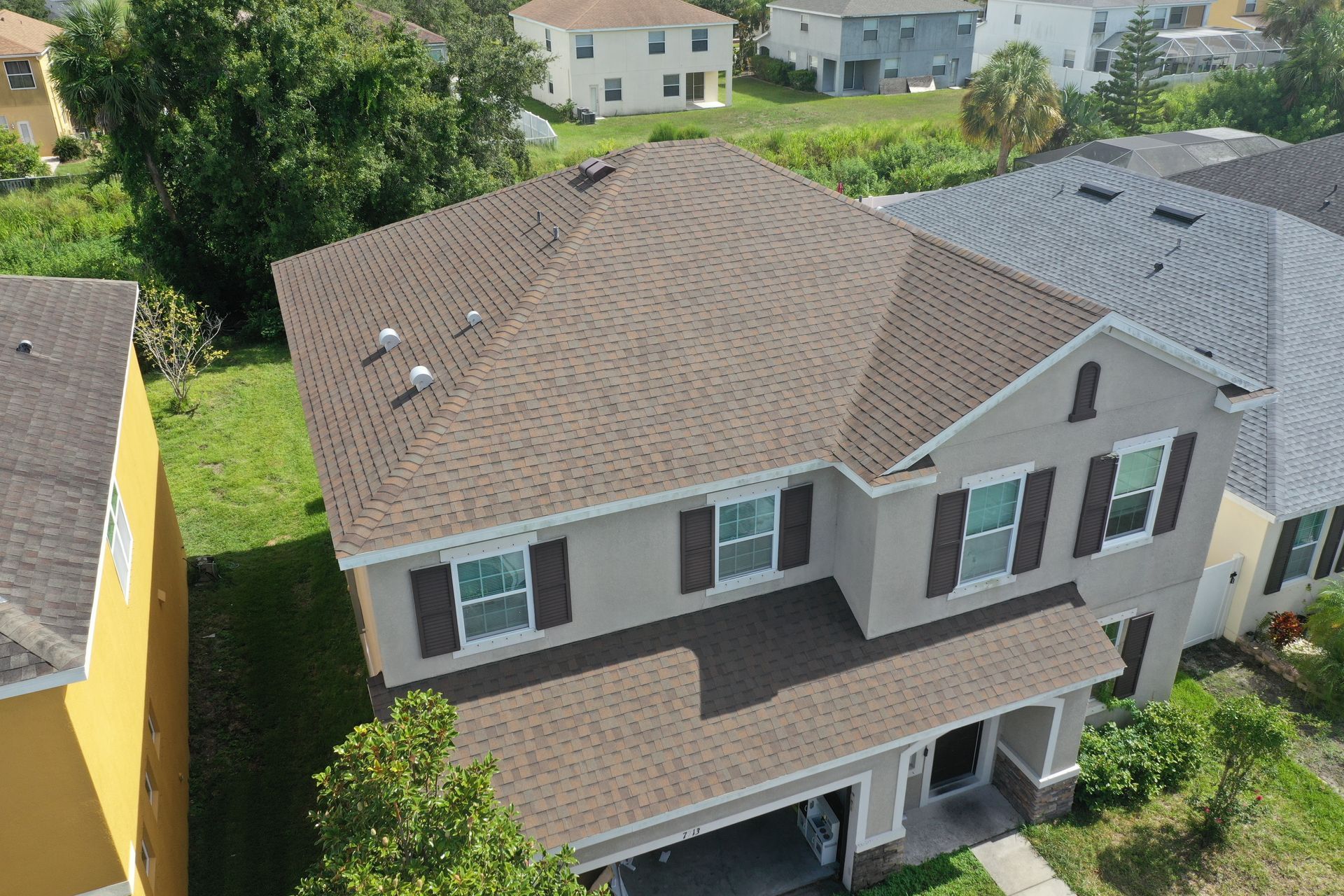 Two-story tan house with brown roof and shutters. Green grass surrounds the home.