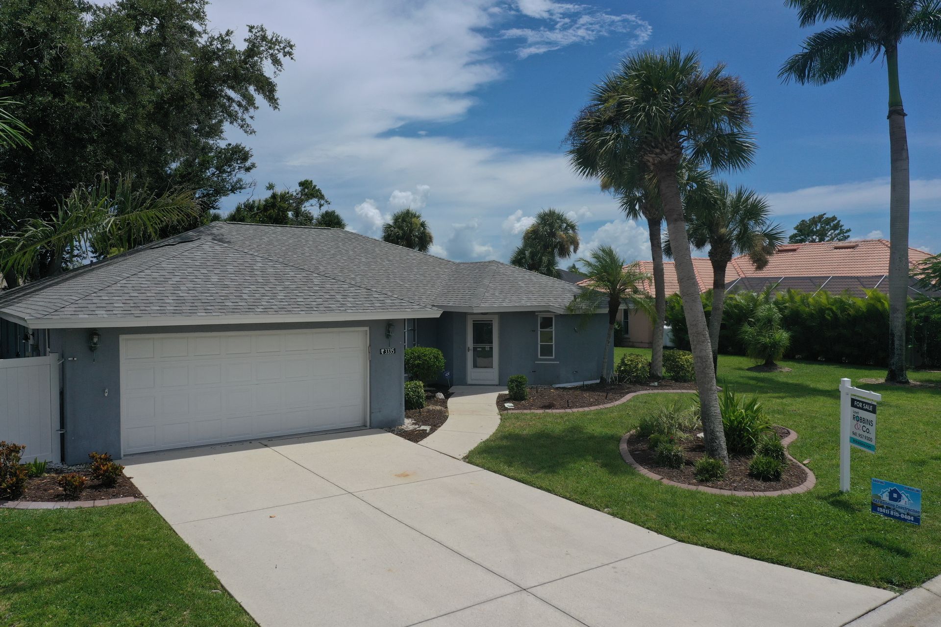 Single-story blue house with a driveway and landscaping on a sunny day, palm trees, and a for sale sign.