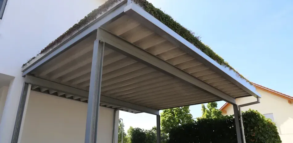 Metal pergola with green roof, over outdoor area of white building. Clear blue sky.