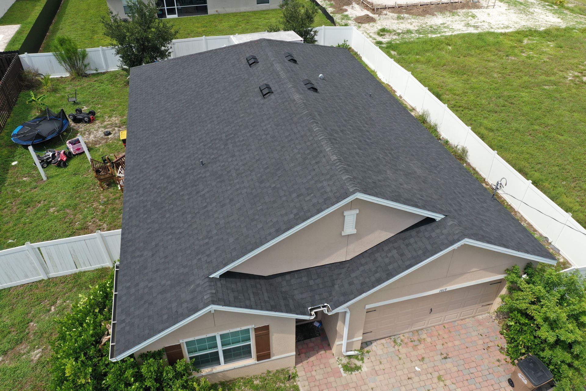 Overhead view of a house with a dark gray roof and tan exterior. Green grass surrounds the house, white fence along the side.