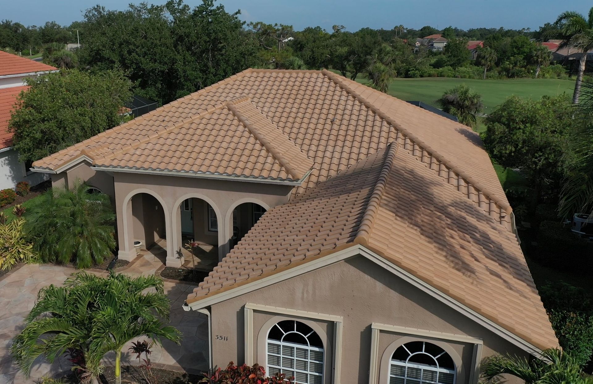 Tan stucco house with arched entry, brown tile roof, and surrounding trees.