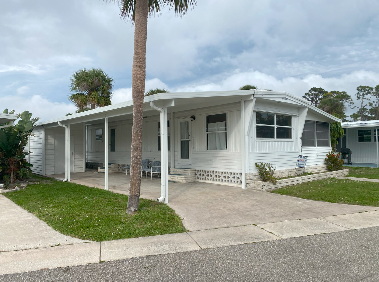 White mobile home with carport, palm tree, and driveway on a sunny day.