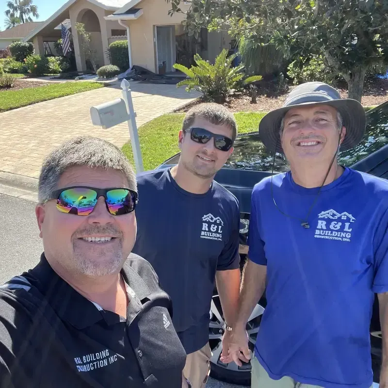 Three men in front of a house. One wearing sunglasses, one wearing a hat, all smiling, in blue and black shirts.