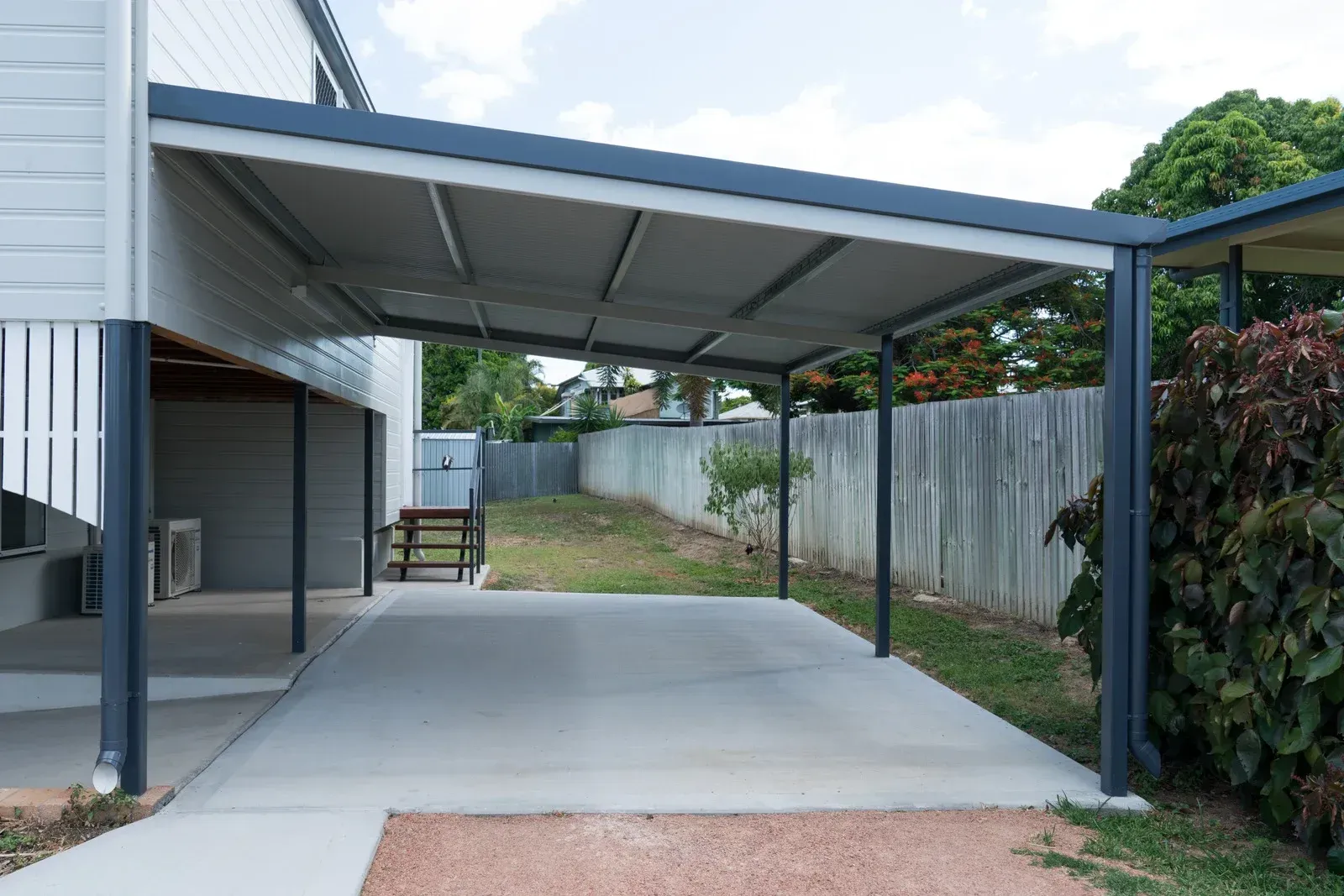 Carport attached to a light-colored house with gray roof and concrete driveway, bordered by a yard and fence.