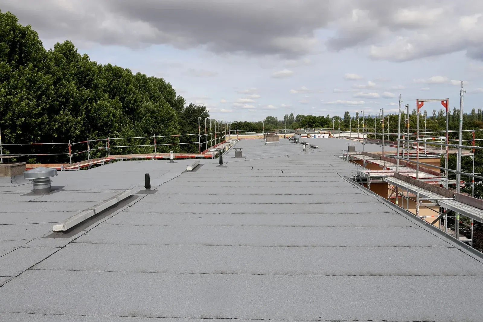 A wide, gray, flat roof with some vents and scaffolding under a cloudy sky.