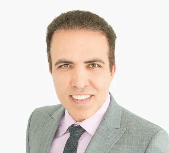 Smiling man with dark hair in a grey suit and tie, light pink shirt against a white background.